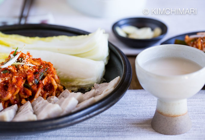 korean pork belly, spicy radish salad and cabbages served on plate with a glass of makgeolli on the side