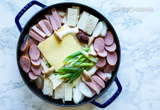 Pot filled with sliced kimchi, spam, tofu, sausage, tteok topped with cheese and gochujang